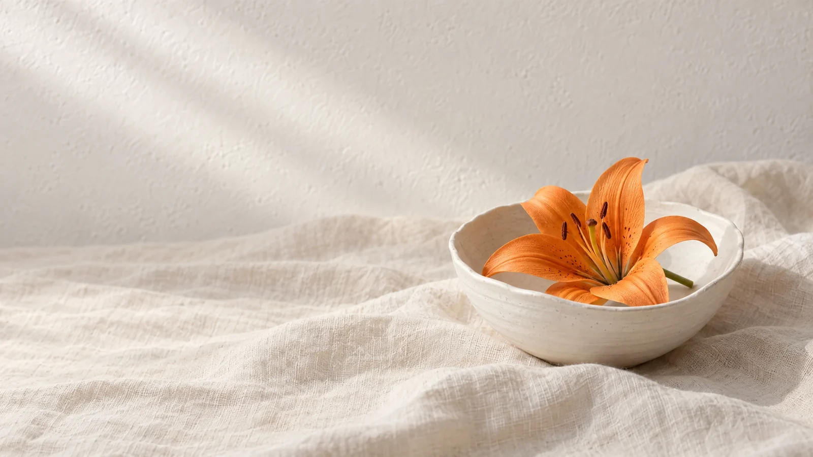 Matte, handgeformte weiße Schale mit einer orangefarbenen, fein gesprenkelten Lilienblüte und kurzem grünem Stielansatz auf zerknittertem cremefarbenem Leinen vor warm-weißer, strukturierter Putzwand mit zarten diagonalen Lichtstreifen von links oben